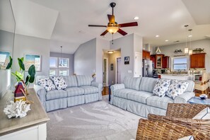 A spacious living room with a large ceiling fan, two sofas with patterned cushions, and a kitchen area visible through an open doorway. - Paws in the Sand -- Gorgeous Pet Friendly Home with Pool (Kitty Hawk)