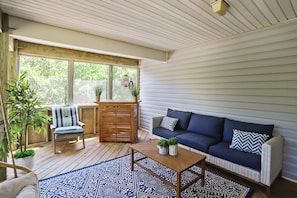 A sunlit room with a wooden floor, a blue patterned rug, a sofa with cushions, a chair, and a small table with plants. - Ola Corolla -- Pet Friendly Oceanside home with Pool, Hot Tub (Corolla)