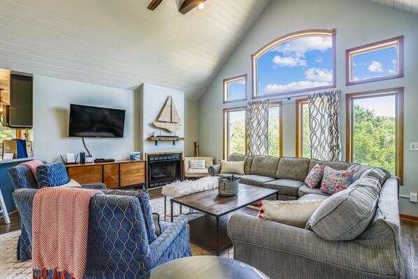 A spacious living room with a fireplace, a television, and a large window with curtains. - Ola Corolla -- Pet Friendly Oceanside home with Pool, Hot Tub (Corolla)