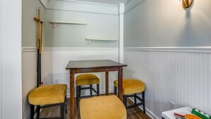 A small dining area with a table and four chairs, a wall-mounted light fixture, and a shelf with a broom. - Ola Corolla -- Pet Friendly Oceanside home with Pool, Hot Tub (Corolla)