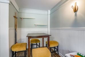 Um pequeno espaço de jantar com uma mesa e quatro cadeiras, um candeeiro fixado na parede e uma prateleira com uma vassoura.