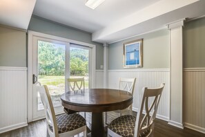 A dining area with a round table and four chairs, a sliding glass door leading to a garden, and a framed picture on the wall. - Ola Corolla -- Pet Friendly Oceanside home with Pool, Hot Tub (Corolla)