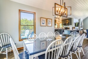 A dining area with a glass table and white chairs, a kitchen with modern appliances, and a large window with a view of trees. - Ola Corolla -- Pet Friendly Oceanside home with Pool, Hot Tub (Corolla)