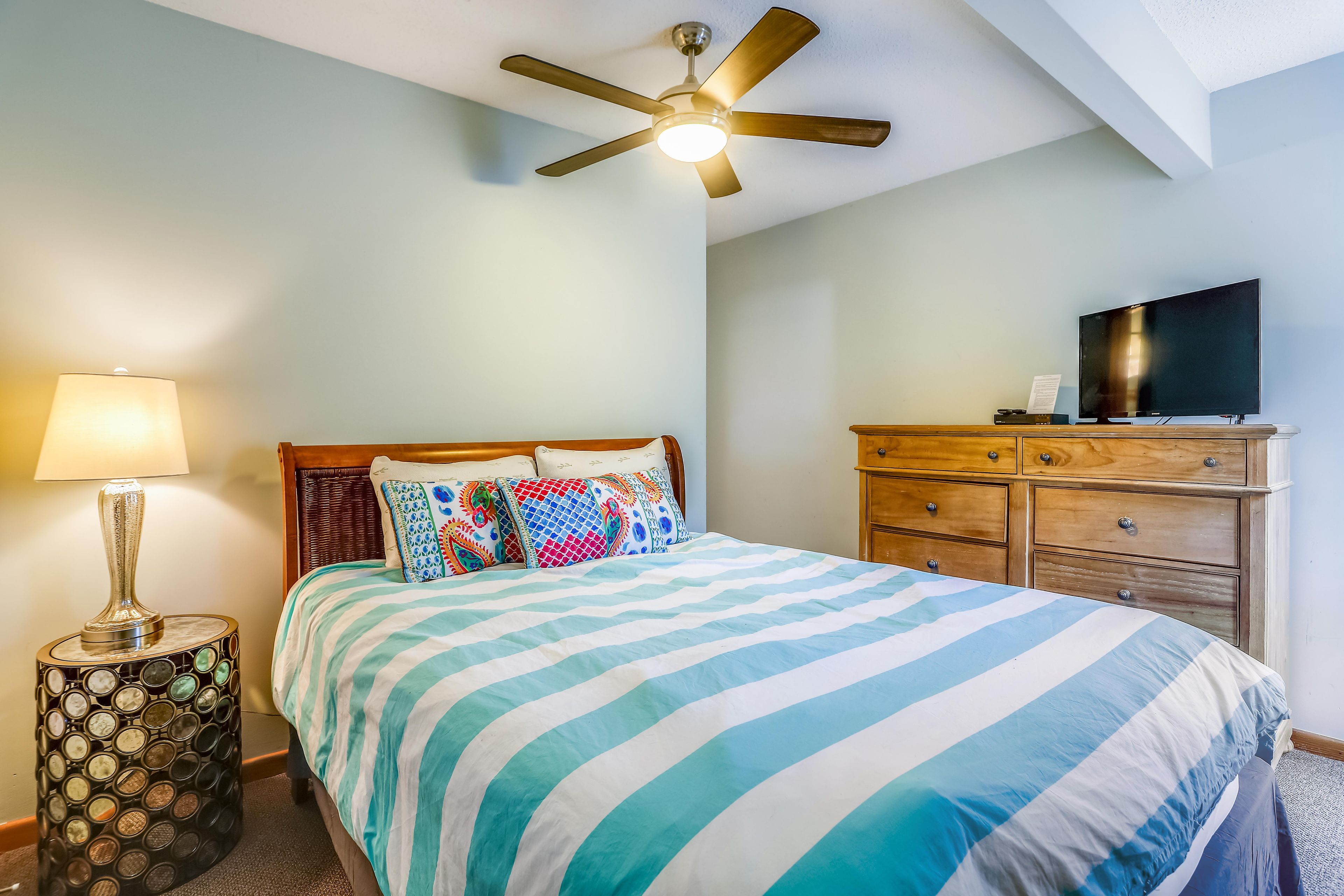 A bedroom with a bed, a ceiling fan, a lamp, a dresser, and a television.