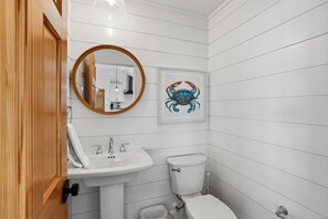 Une salle de bain avec une porte en bois, un lavabo blanc, des toilettes, un miroir avec un cadre en bois et une œuvre d’art représentant un crabe, encadrée.