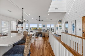 A spacious kitchen with a central island, a dining area with wicker chairs, a living room with a sofa and a view of the ocean, and a staircase leading to an upper level. - Knot for Sail -- Newer Soundfront Home with Hot Tub and Firepit (Duck)