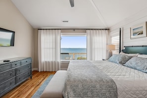 A bedroom with a bed, a dresser, a window with curtains, a ceiling fan, and a view of the sea.