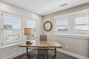 A room with a glass-top table, a woven chair, a wall clock, and white blinds on the windows. - Knot for Sail -- Newer Soundfront Home with Hot Tub and Firepit (Duck)