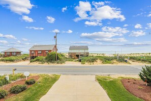 Una carretera costera con acera que conduce a propiedades frente a la playa, incluyendo un edificio rojo y una estructura verde con terraza de madera.
