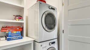Eine übereinander gestellte Waschmaschine und ein Trockner in einem Waschraum, über dem Regale mit Gesellschaftsspielen angebracht sind.