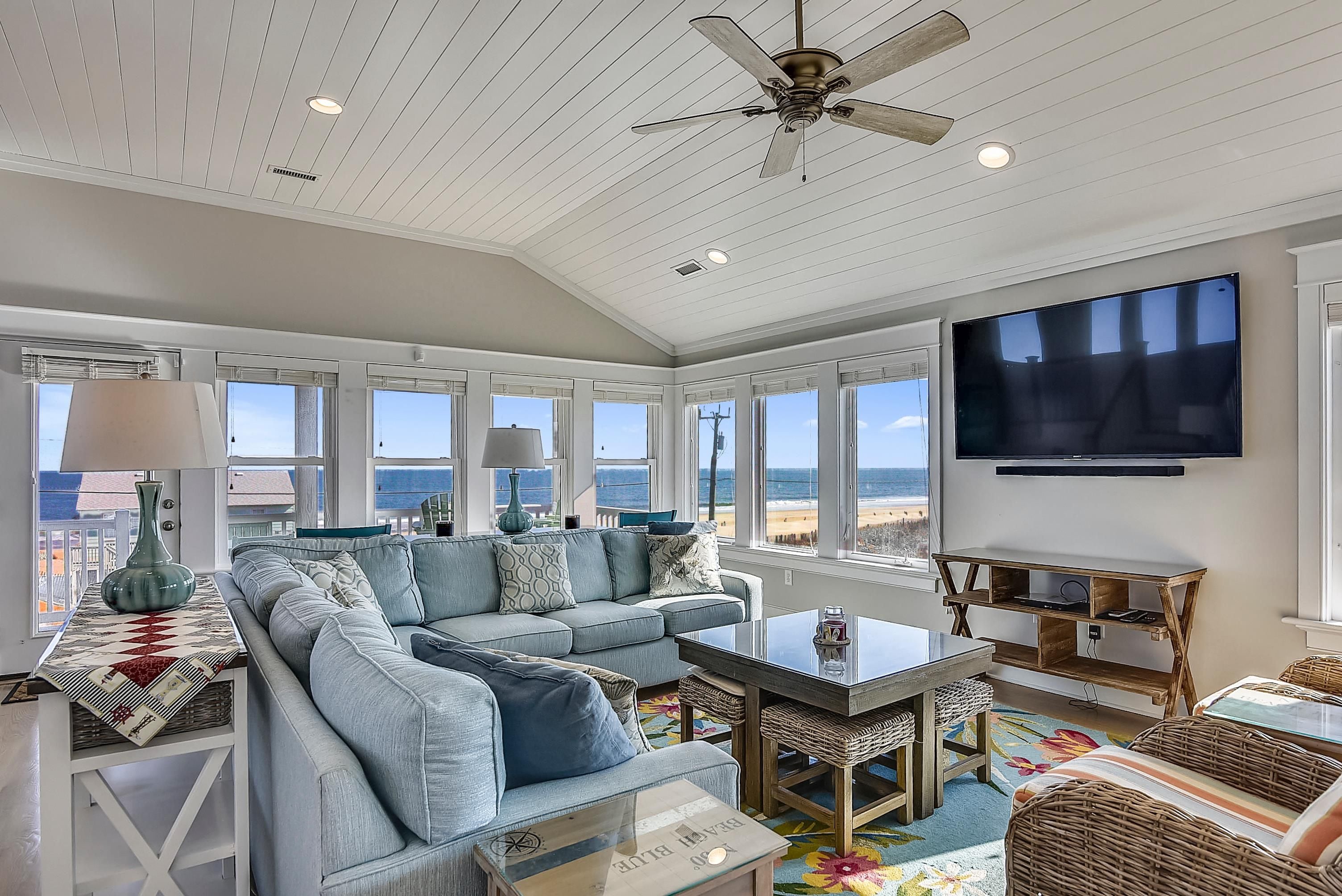 A spacious living room with a large sofa, a television mounted on the wall, and a ceiling fan.