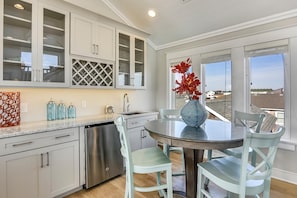 A modern kitchen with a dining area, featuring a stainless steel dishwasher, a round glass table, and light blue chairs. - Fantasea -- Spectacular Semi OF Home in Kitty Hawk w/Elevator, Pool and Ocean Views (Kitty Hawk)
