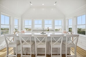 A dining area with a white table and chairs, large windows with a view of the sea, and a high ceiling with recessed lighting.