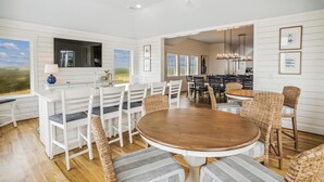 Un vaste espace de salle à manger avec une table ronde en bois entourée de chaises blanches et de poufs en osier, une grande fenêtre offrant une vue sur l’extérieur et un coin cuisine visible à travers une porte ouverte.