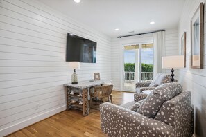 A room with a wooden floor, a dining table with chairs, a sofa, a TV mounted on the wall, and a balcony with a view. - Newbern - New Cons. Semi Oceanfront in Corolla with Fire Pit, Elevator, Hot Tub and More! (Corolla)
