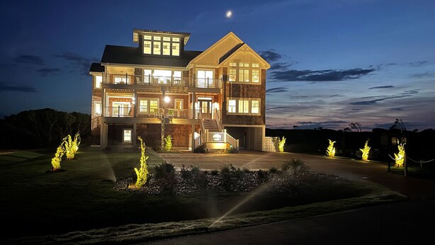 A large, multi-story house with a prominent balcony and multiple windows, illuminated at night. - Newbern - New Cons. Semi Oceanfront in Corolla with Fire Pit, Elevator, Hot Tub and More! (Corolla)