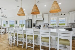 A modern kitchen with white cabinetry, a central island with bar stools, and large windows offering a view of the outdoors. - Newbern - New Cons. Semi Oceanfront in Corolla with Fire Pit, Elevator, Hot Tub and More! (Corolla)