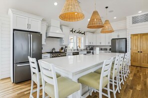 A modern kitchen with white cabinetry, stainless steel appliances, a central island with bar seating, and a herringbone-patterned backsplash. - Newbern - New Cons. Semi Oceanfront in Corolla with Fire Pit, Elevator, Hot Tub and More! (Corolla)