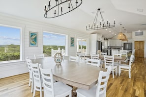 A spacious dining area with a large wooden table and white chairs, a chandelier, and artwork on the walls. - Newbern - New Cons. Semi Oceanfront in Corolla with Fire Pit, Elevator, Hot Tub and More! (Corolla)