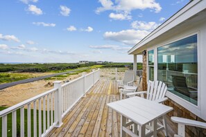 A wooden deck with white railings, a table, and chairs, overlooking a beach and distant buildings. - Newbern - New Cons. Semi Oceanfront in Corolla with Fire Pit, Elevator, Hot Tub and More! (Corolla)