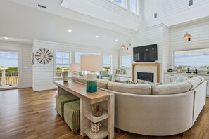 A spacious living room with a large sofa, a wooden coffee table, and a television mounted on the wall. - Newbern - New Cons. Semi Oceanfront in Corolla with Fire Pit, Elevator, Hot Tub and More! (Corolla)