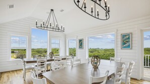 A spacious dining area with a large table and chairs, large windows with a view of greenery, and a chandelier. - Newbern - New Cons. Semi Oceanfront in Corolla with Fire Pit, Elevator, Hot Tub and More! (Corolla)