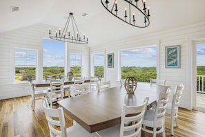 A spacious dining area with a large table and chairs, large windows with a view of greenery, and a chandelier. - Newbern - New Cons. Semi Oceanfront in Corolla with Fire Pit, Elevator, Hot Tub and More! (Corolla)