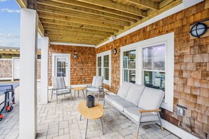 A covered patio area with a wooden ceiling, two windows, a sofa, a chair, and a small table. - Newbern - New Cons. Semi Oceanfront in Corolla with Fire Pit, Elevator, Hot Tub and More! (Corolla)