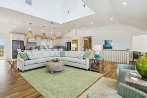 A spacious living area with a large L-shaped sectional sofa, a round woven ottoman, and a dining area with a table and chairs. - Newbern - New Cons. Semi Oceanfront in Corolla with Fire Pit, Elevator, Hot Tub and More! (Corolla)