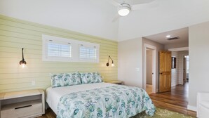 A bedroom with a bed, a nightstand, a window with blinds, a ceiling fan, and a door leading to another room. - Newbern - New Cons. Semi Oceanfront in Corolla with Fire Pit, Elevator, Hot Tub and More! (Corolla)