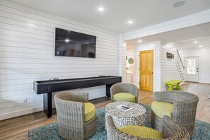 A modern living room with a flat-screen TV mounted on the wall, a black console table, wicker chairs with cushions, and a wooden door leading to another room. - Newbern - New Cons. Semi Oceanfront in Corolla with Fire Pit, Elevator, Hot Tub and More! (Corolla)