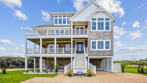A two-story house with a balcony, a front porch, and a sign for Atlantic Realty. - Newbern - New Cons. Semi Oceanfront in Corolla with Fire Pit, Elevator, Hot Tub and More! (Corolla)