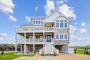 A two-story house with a balcony, a front porch, and a sign for Atlantic Realty. - Newbern - New Cons. Semi Oceanfront in Corolla with Fire Pit, Elevator, Hot Tub and More! (Corolla)