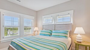 A bedroom with a large bed, two bedside lamps, and a view of greenery through the windows. - Newbern - New Cons. Semi Oceanfront in Corolla with Fire Pit, Elevator, Hot Tub and More! (Corolla)