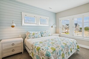A bedroom with a bed, nightstands, a window with blinds, and a view of a building outside. - Newbern - New Cons. Semi Oceanfront in Corolla with Fire Pit, Elevator, Hot Tub and More! (Corolla)