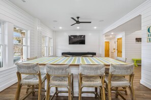 A dining area with a striped table, four wicker chairs, a ceiling fan, and a TV mounted on the wall. - Newbern - New Cons. Semi Oceanfront in Corolla with Fire Pit, Elevator, Hot Tub and More! (Corolla)