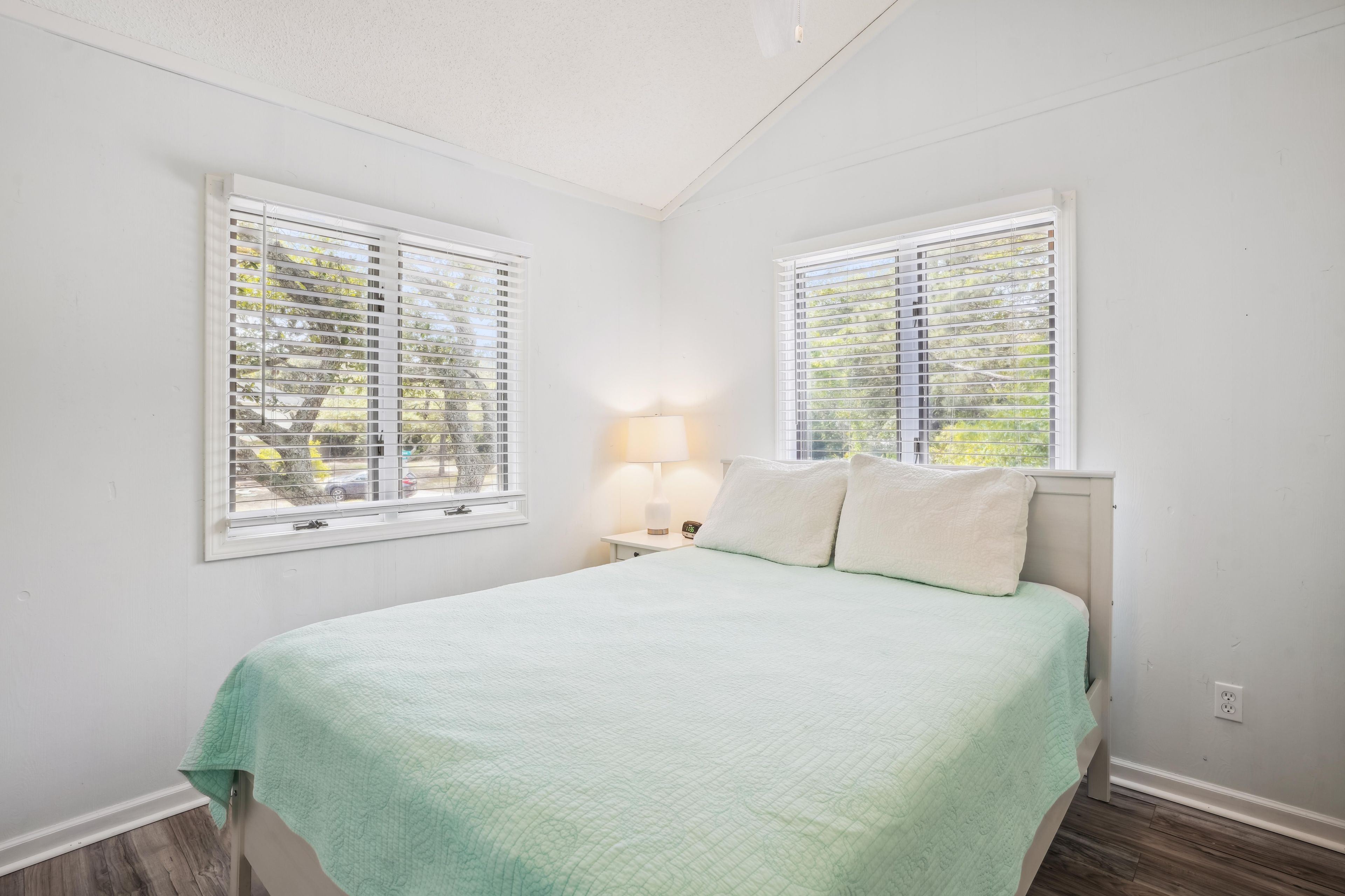 A bedroom with a bed, two windows, a nightstand, and a lamp.