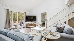 En moderne stue med sofa, sofabord og fjernsyn. En trappe til højre og en balkon med udsigt til træer.