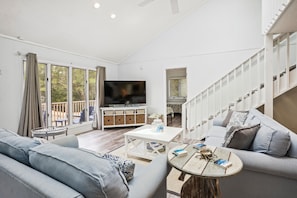 Ein modernes Wohnzimmer mit einer Couch, einem Couchtisch und einem Fernseher. Eine Treppe befindet sich rechts, und ein Balkon bietet Blick auf Bäume.