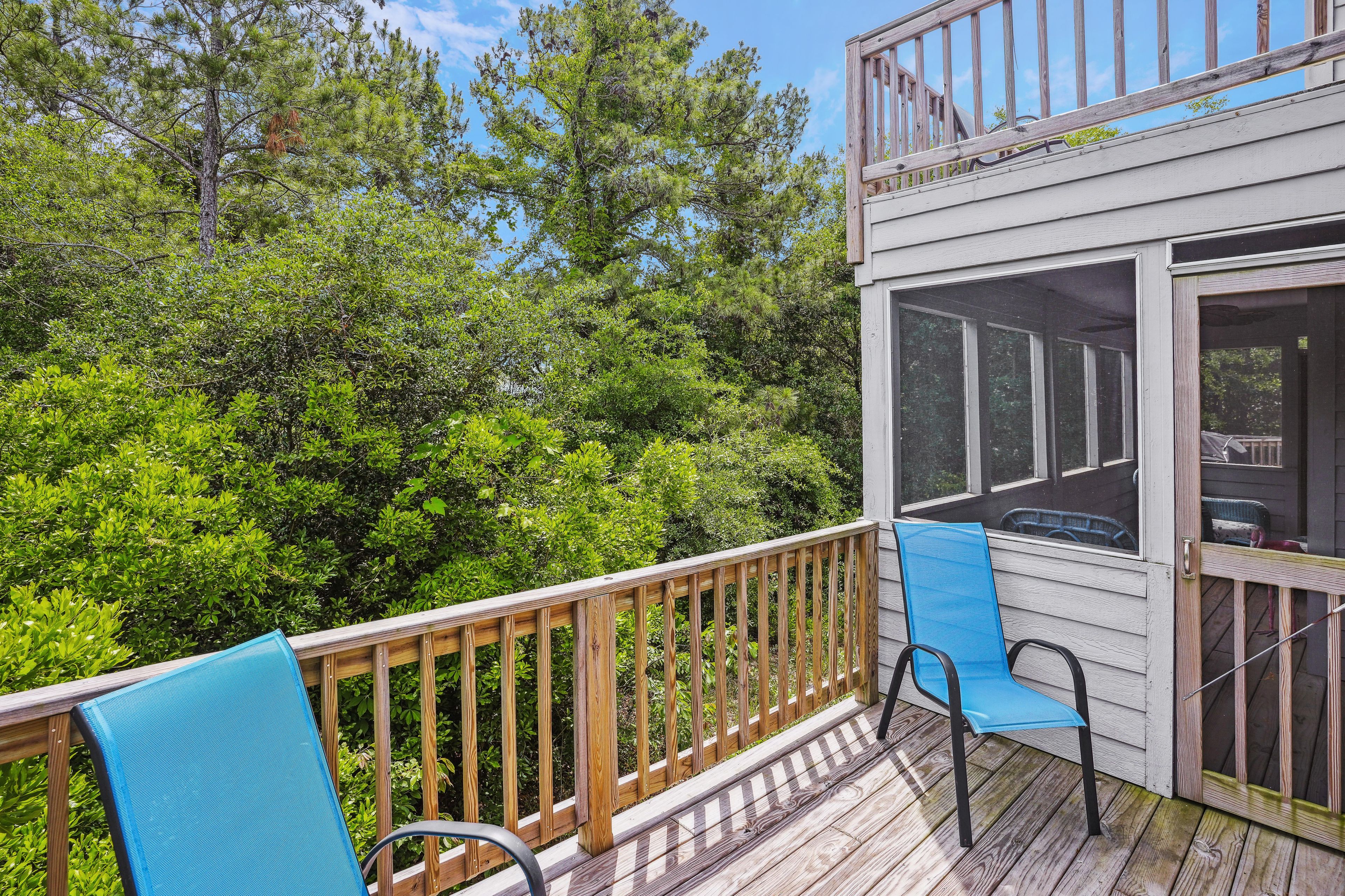 A wooden deck with a railing, two blue chairs, and a view of dense green foliage.