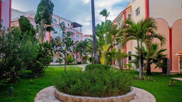Une cour dotée d’un chemin circulaire pavé de pierres, bordée d’une végétation luxuriante et de palmiers, qui mène à un bâtiment aux murs extérieurs de couleur rose et beige.
