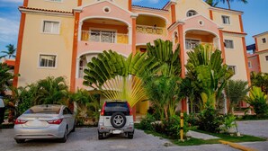 Un hôtel à plusieurs étages, doté de balcons, de palmiers et d’une voiture garée.