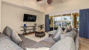 A modern living room with a large sectional sofa, a flat-screen TV, a coffee table, and a view of the outdoors through large windows.