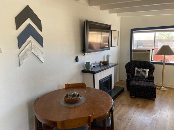 A dining area with a round wooden table and chairs, a fireplace, a television mounted above it, and a patterned armchair by a window. - Apartment in downtown Joshua Tree - short walk to the restaurants and shops (Joshua Tree)