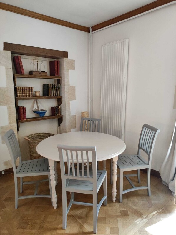 A dining area with a round table and four chairs, a bookshelf with books and decorative items, and a window with blinds. - Chambre D'hôte à La Cour Souveraine Près des Plages du Débarquement et de Bayeux (Mosles)
