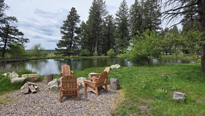 A lakeside setting with two Adirondack chairs on a pebbled area, surrounded by trees and a calm lake.