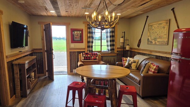 A cozy interior with a wooden ceiling, a chandelier, a dining table with stools, a sofa with patterned cushions, a red refrigerator, and a television on the wall.