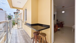 A balcony with a kitchen area, including a counter and stools, overlooking a residential area. - Raridade 2 vagas Praia Grande - Nobre  (Ubatuba)