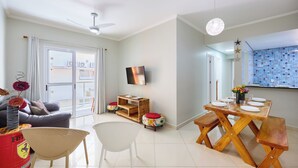 A modern living room with a sofa, a coffee table, a TV, and a dining area with a wooden table and benches. - Raridade 2 vagas Praia Grande - Nobre  (Ubatuba)