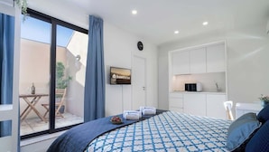 A modern hotel room with a large bed, a sliding glass door leading to a balcony, a small kitchenette, and a mounted television.